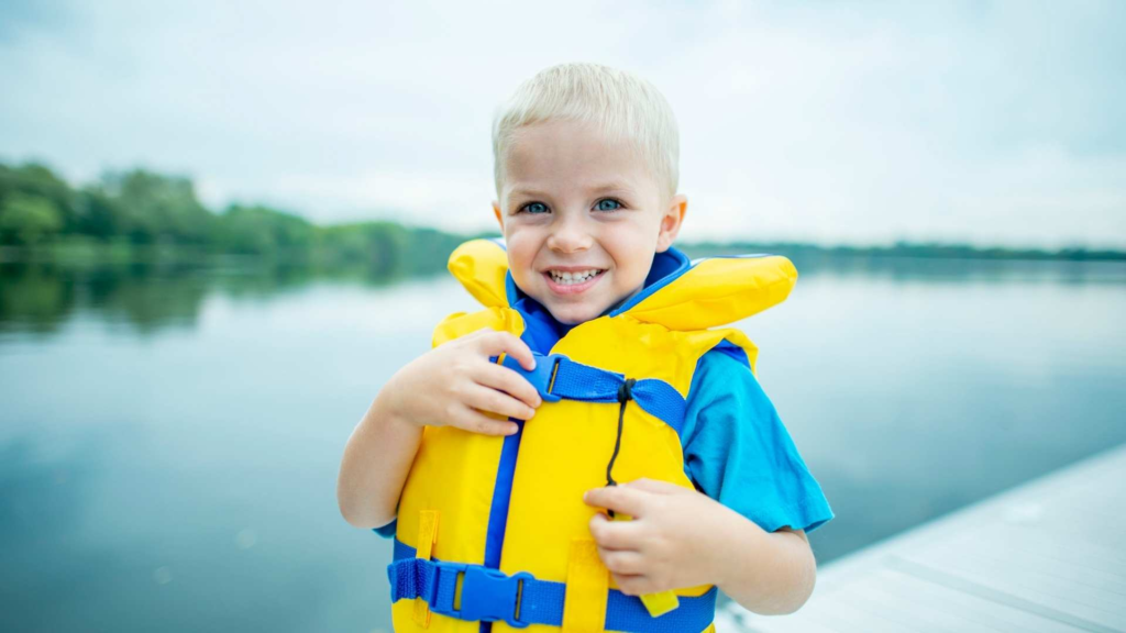 Young child wearing a yellow life jacket by a lake, illustrating summer water safety for families.