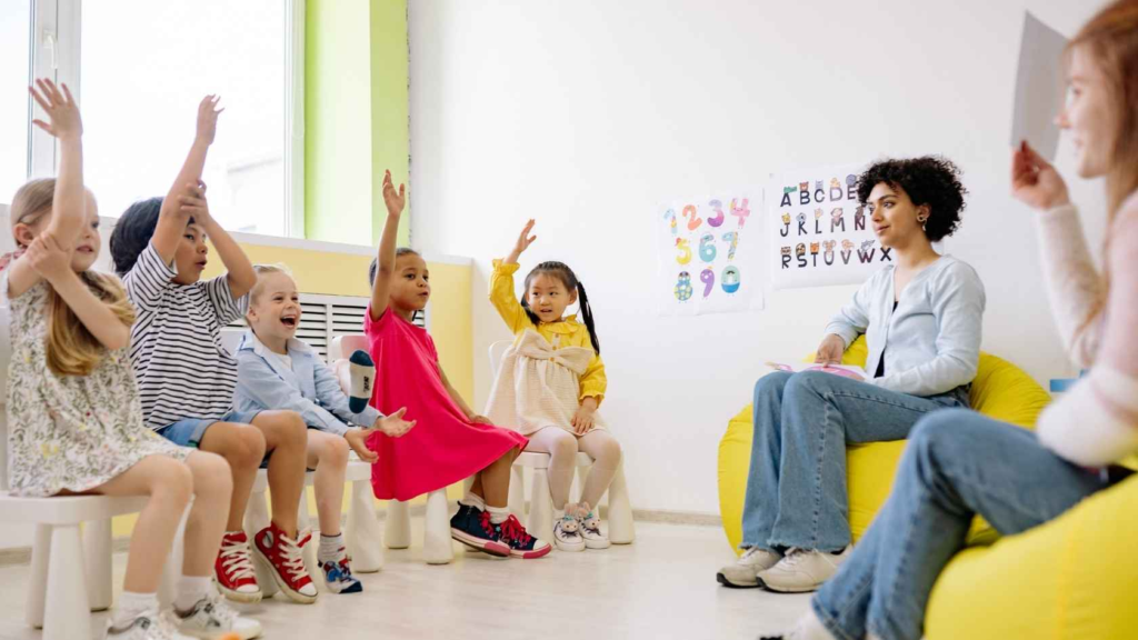 Preschool children raising their hands during a group classroom activity with teachers.