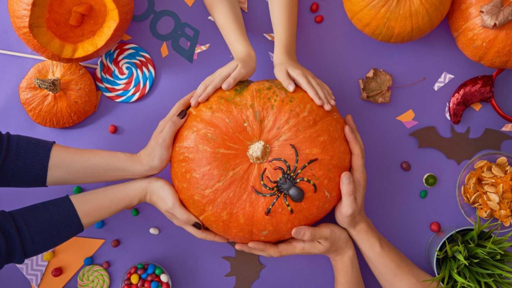 Hands holding a pumpkin surrounded by Halloween decorations, candy, and fall craft materials.
